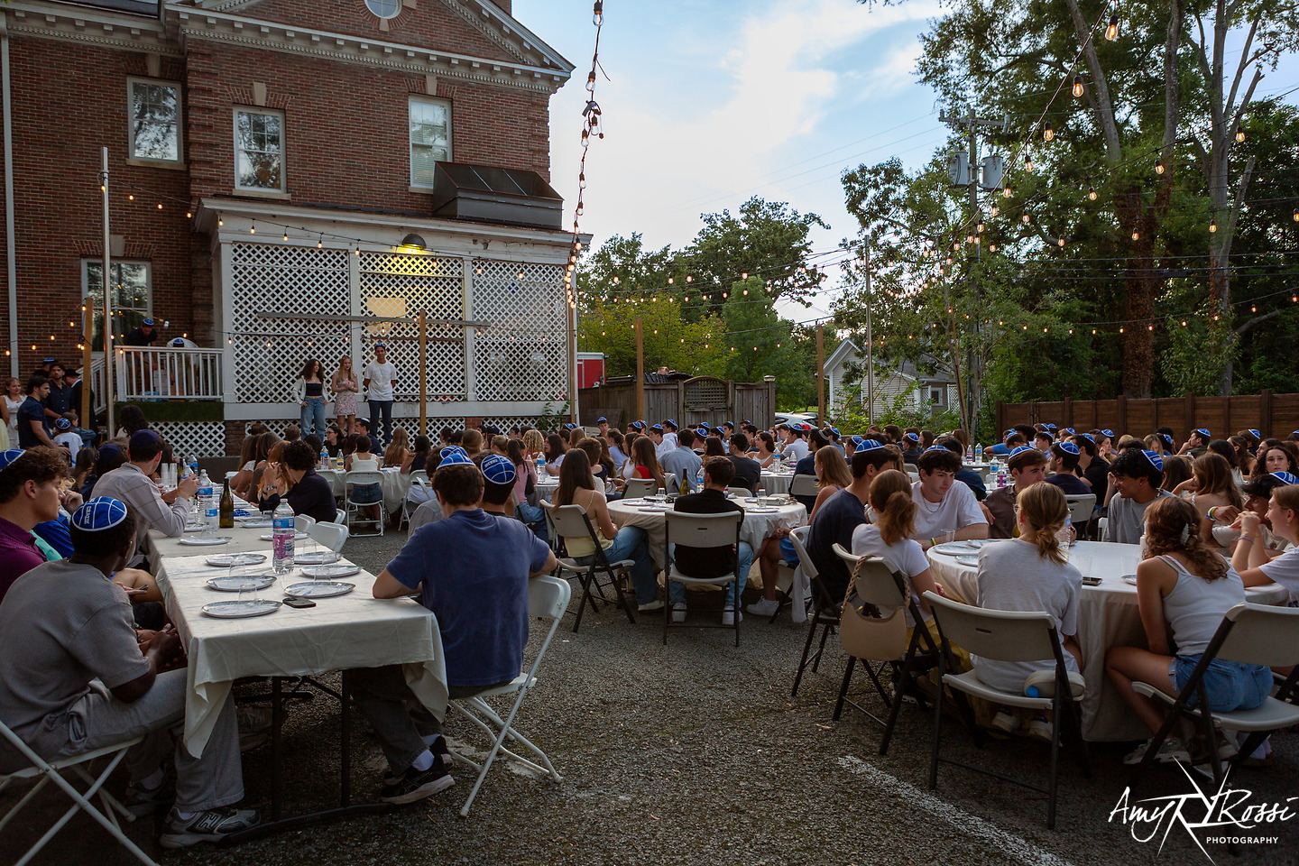 Round tables full of students under string lights at dusk in the backyard